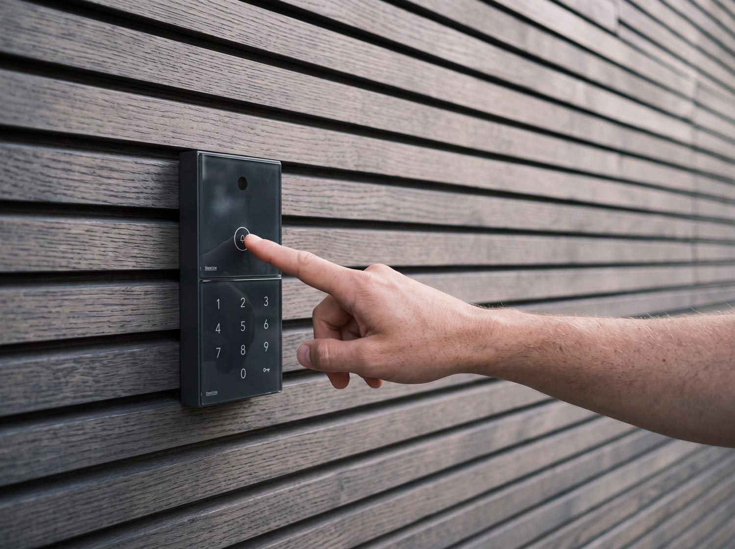 Hand tippt auf ein schwarzes, rechteckiges Türsprechanlagengerät mit Tastenfeld an einer Holzlattenwand