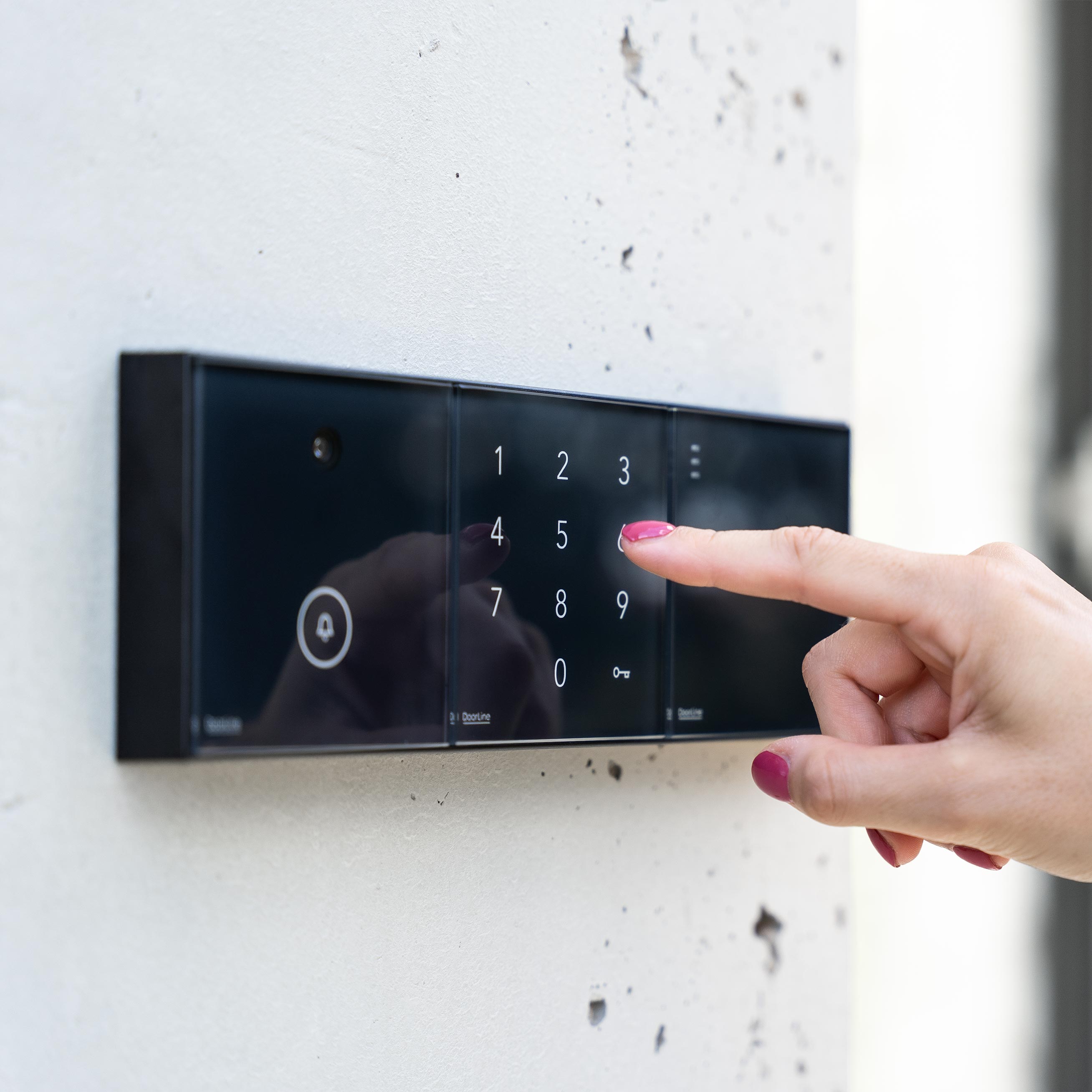 Hand mit pink lackiertem Fingernagel tippt auf ein schwarzes, wandmontiertes Touch-Tastenfeld mit Zahlen von 0 bis 9