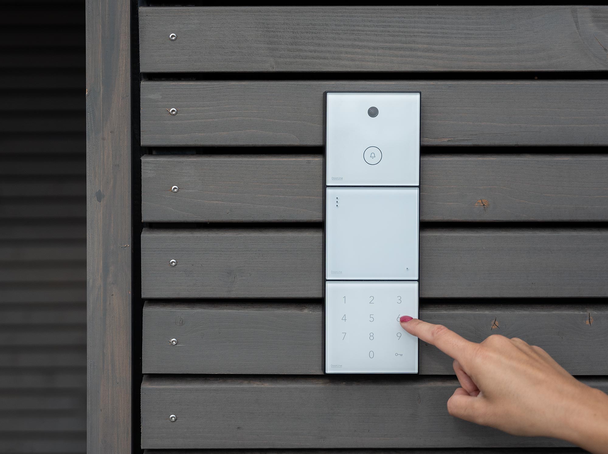 Hand tippt auf ein weißes, modernes Türcodefeld mit Kamera an einer dunkelgrauen Holzfassade