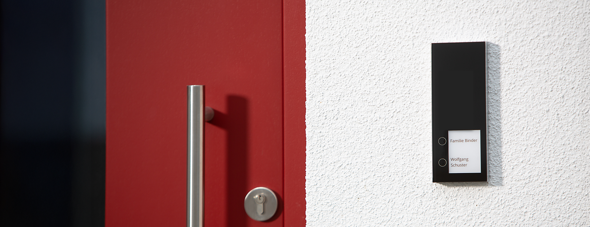 Rote Haustür mit Edelstahlgriff und Schlüsselloch, daneben an weißer Wand eine schwarze DoorLine Türsprechanlage mit zwei Klingeltasten und Namensschildern ‚Familie Binder‘ und ‚Wolfgang Schuster‘.
